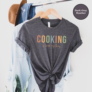 May include: A dark grey heather t-shirt with the text "COOKING is kinda my thing" in colorful letters. The shirt is hanging on a wooden hanger.