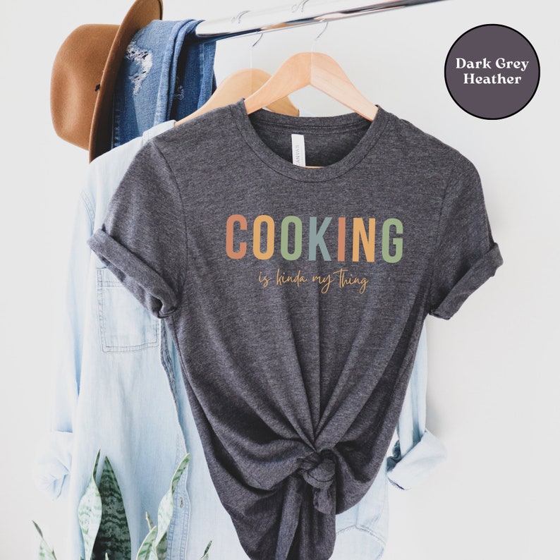 May include: A dark grey heather t-shirt with the text "COOKING is kinda my thing" in colorful letters. The shirt is hanging on a wooden hanger.