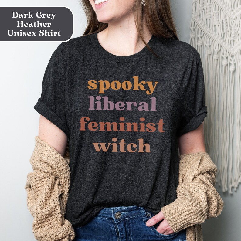 May include: A dark grey heather unisex t-shirt with the text "spooky liberal feminist witch" printed in orange, pink, and brown.