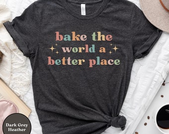May include: Dark grey heather t-shirt with the text "bake the world a better place" in rainbow colors with gold glitter accents.