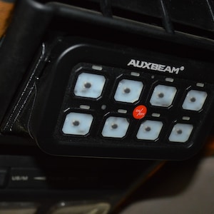 May include: A black control panel with the text "AUXBEAM" and eight buttons. The buttons are arranged in two rows of four. The buttons are labeled with numbers.
