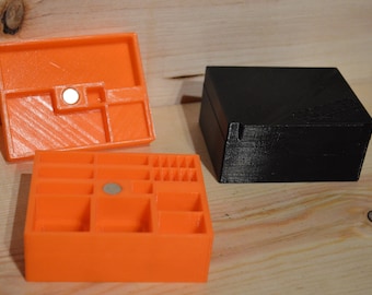 May include: Two 3D printed boxes, one black and one orange. The orange box has a lid and a compartmentalized base. Both boxes have a small magnet on the side.