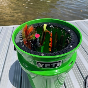 May include: A bright green YETI fishing tackle bucket filled with various fishing lures. The lures are suspended from a black ring inside the bucket. The bucket is sitting on a dock with water in the background.