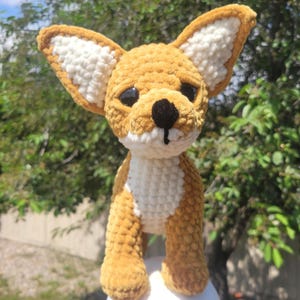 May include: A handmade crochet Chihuahua dog with a tan body, white chest, and large, upright ears. The dog has a black nose and eyes, and is standing on a white surface. The plush toy is a charming example of crochet art.
