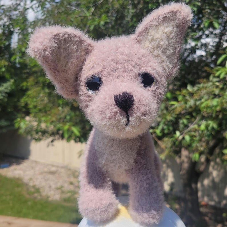 May include: A handmade plush toy dog in a soft mauve color. The dog has large, floppy ears, a black nose, and black eyes. The plushie is made of a fuzzy material, giving it a cuddly appearance. The background is blurred, with green foliage.