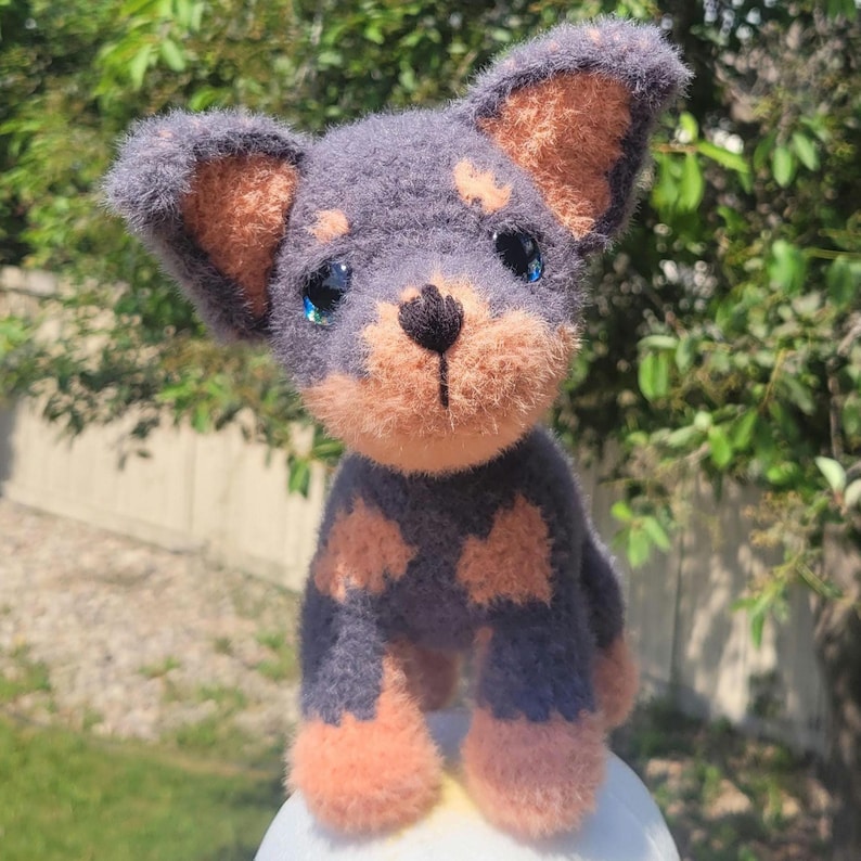 May include: A handmade crochet dog figurine with a gray body, tan markings, and large blue eyes. The dog has tan ears, paws, and muzzle. The plush toy is standing on a white surface, with a green background.