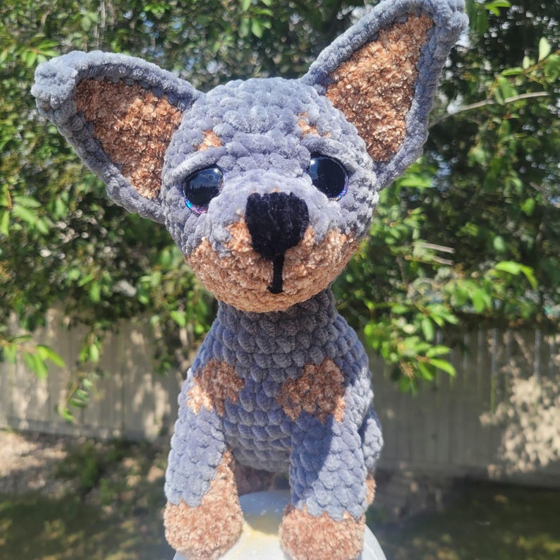 May include: A handmade crochet dog in a seated position. The plush toy features a gray body with brown accents on the ears, paws, and spots. It has large, expressive eyes and a black nose. The dog is made of soft, textured yarn.