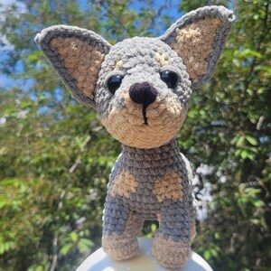 May include: A handmade crochet chihuahua dog in gray, tan, and black. The dog has large, upright ears, a black nose, and black eyes. The crochet dog is standing on a white surface against a blurred green background.