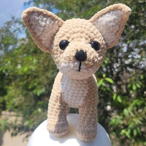 May include: A crocheted chihuahua dog figurine in tan and cream colors. The dog has large, upright ears, black eyes, and a black nose. The handmade dog is standing on a white surface, with a blurred green background.