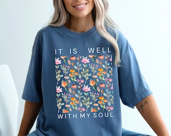 May include: A blue t-shirt with the words "IT IS WELL WITH MY SOUL" in white. The shirt features a floral design in a rectangular shape, with pink, orange, and yellow flowers and green leaves. The shirt is a relaxed fit.