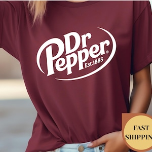 May include: Burgundy t-shirt featuring the Dr Pepper logo in white. The logo includes the brand name and "Est. 1885" in a curved design. The shirt is a short-sleeved crew neck.