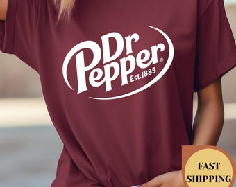 Może przedstawiać: Bordowa koszulka z białym logo Dr Pepper. Logo zawiera nazwę marki i napis "Est. 1885" w zakrzywionym kształcie. Koszulka ma krótki rękaw i okrągły dekolt.