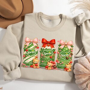 May include: A beige sweatshirt featuring three green Sprite soda cans decorated with bows, hearts, and teddy bears. The cans have a heart-shaped design and are adorned with cherries. The sweatshirt has long sleeves and a crew neck.