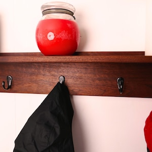 May include: A dark wooden shelf with three metal hooks. A large red jar with a glass lid sits on the shelf. A black jacket hangs from one hook. A red hat is partially visible on the right.