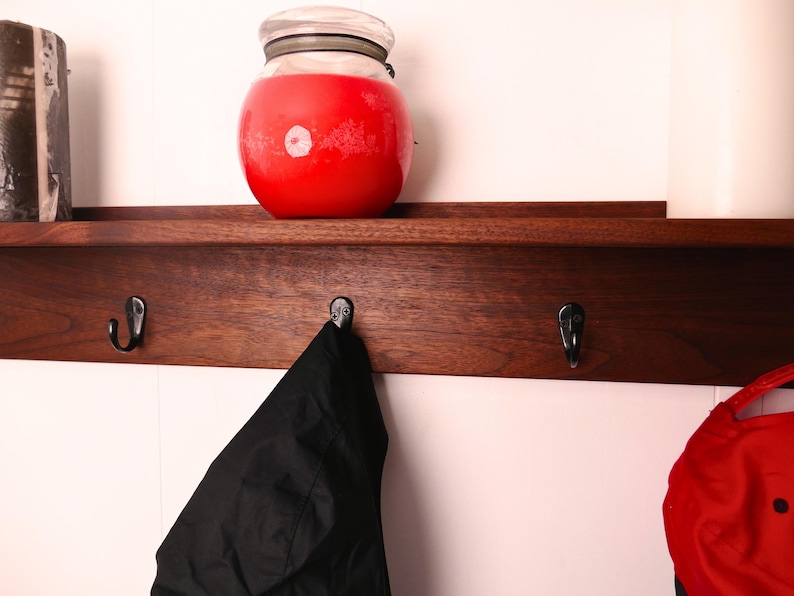 May include: A dark wooden shelf with three metal hooks. A large red jar with a glass lid sits on the shelf. A black jacket hangs from one hook. A red hat is partially visible on the right.