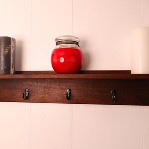 May include: A dark wood shelf with five black hooks, holding a black and gray candle, a red jar with a lid, and a white candle. The shelf is mounted on a white wall.