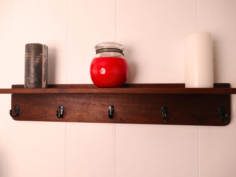 May include: A dark wood shelf with five black hooks, holding a black and gray candle, a red jar with a lid, and a white candle. The shelf is mounted on a white wall.