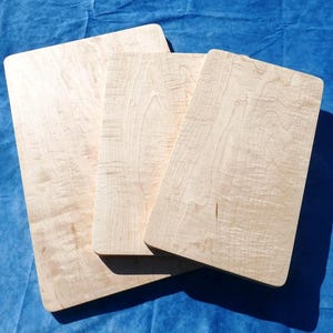 May include: Three light-colored wooden cutting boards with rounded corners are stacked on a blue surface. The wood grain is visible, and the boards vary slightly in size. The boards are rectangular.