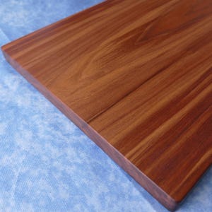 May include: A rectangular wooden cutting board with a rich, reddish-brown finish. The wood grain is visible, and the board has a smooth, polished surface. The board is set against a blue background.