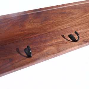 May include: A wooden shelf with a rich, dark brown finish. The shelf features three black metal hooks, perfect for hanging coats, hats, or other items. The wood grain is visible, adding to the natural aesthetic.