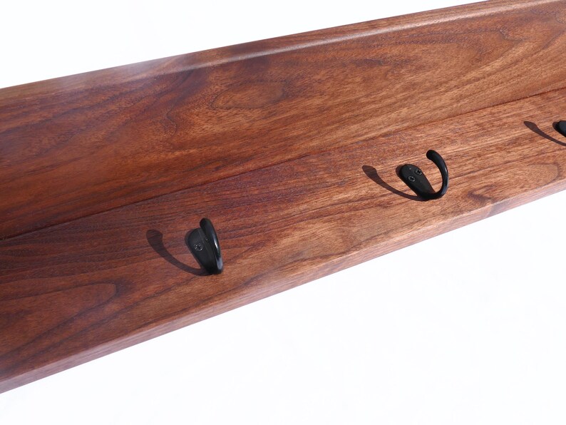 May include: A wooden shelf with a rich, dark brown finish. The shelf features three black metal hooks, perfect for hanging coats, hats, or other items. The wood grain is visible, adding to the natural aesthetic.