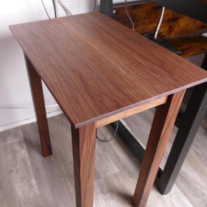 May include: A small, rectangular wooden side table with a rich, dark brown finish. The table has four straight legs and a simple, minimalist design. The tabletop measures approximately 24 inches long and 12 inches wide.