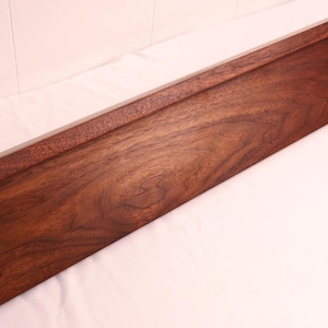 May include: A long, rectangular wooden tray with a rich, dark brown finish. The tray has a raised edge along one side, creating a subtle border. The wood grain is visible, adding texture and warmth to the piece. The tray is likely for serving or display.