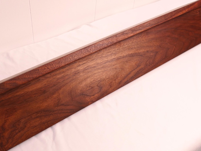 May include: A long, rectangular wooden tray with a rich, dark brown finish. The tray has a raised edge along one side, creating a subtle border. The wood grain is visible, adding texture and warmth to the piece. The tray is likely for serving or display.