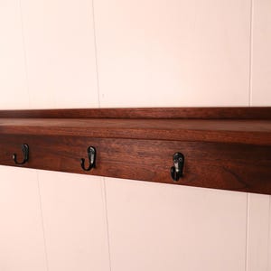May include: A dark brown wooden shelf with five black metal hooks. The shelf has a simple, rectangular design and is mounted on a white wall. The hooks are evenly spaced along the bottom edge of the shelf.