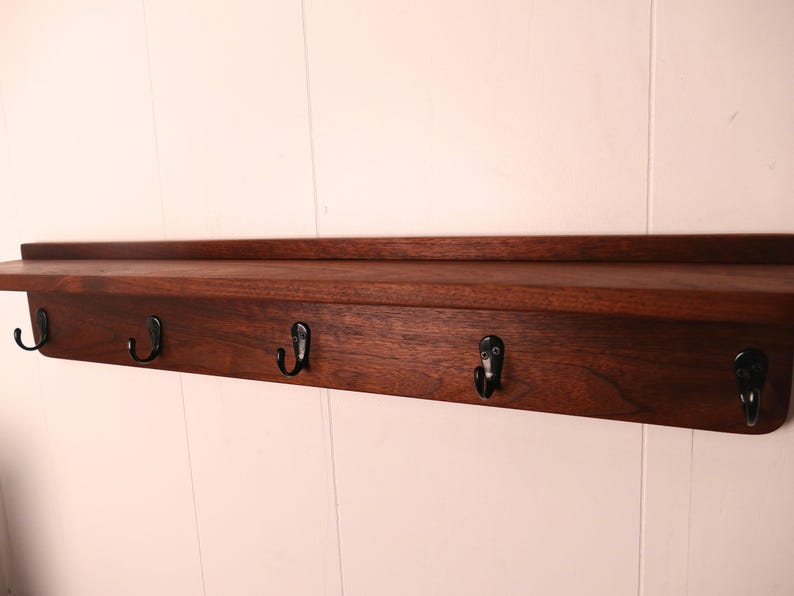 May include: A dark brown wooden shelf with five black metal hooks. The shelf has a simple, rectangular design and is mounted on a white wall. The hooks are evenly spaced along the bottom edge of the shelf.