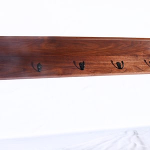 May include: A wooden shelf with a dark brown finish and five black metal hooks. The shelf has a simple, modern design with a slightly angled top edge. Ideal for entryways or bedrooms.