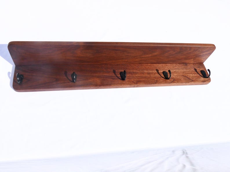 May include: A wooden shelf with a dark brown finish and five black metal hooks. The shelf has a simple, modern design with a slightly angled top edge. Ideal for entryways or bedrooms.
