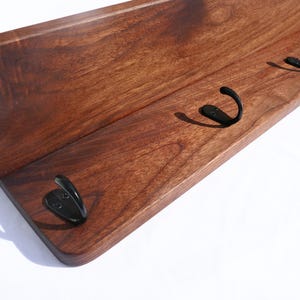 May include: A wooden wall shelf with a dark brown finish. The shelf has a curved front and features four black metal hooks for hanging items. The wood grain is visible, adding texture to the design. The shelf is designed for wall mounting.