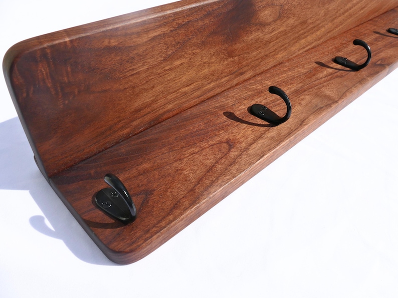 May include: A wooden wall shelf with a dark brown finish. The shelf has a curved front and features four black metal hooks for hanging items. The wood grain is visible, adding texture to the design. The shelf is designed for wall mounting.