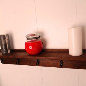 May include: A dark stained wooden shelf with four black hooks, holding a black candle, a red jar candle, and a white pillar candle. The shelf is mounted on a white wall.