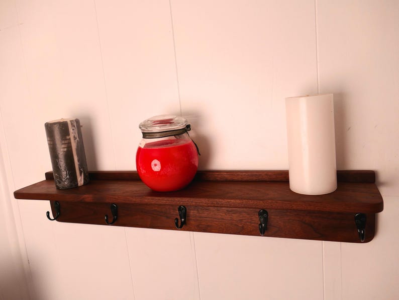 May include: A dark stained wooden shelf with four black hooks, holding a black candle, a red jar candle, and a white pillar candle. The shelf is mounted on a white wall.