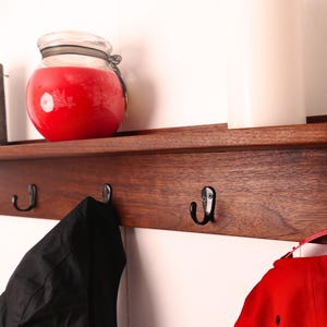 May include: A wooden wall shelf with four black metal hooks. A red candle in a glass jar and a dark brown candle sit on the shelf. A black jacket and a red cap hang from the hooks.