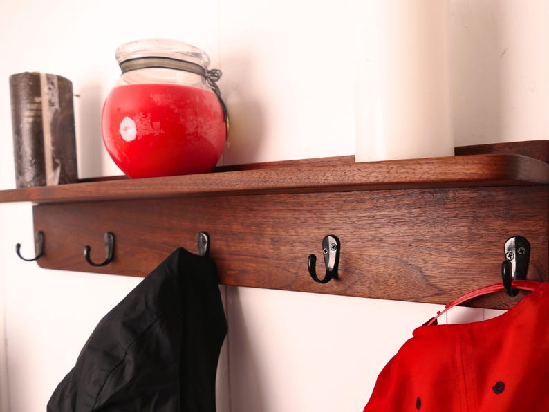 May include: A wooden wall shelf with four black metal hooks. A red candle in a glass jar and a dark brown candle sit on the shelf. A black jacket and a red cap hang from the hooks.