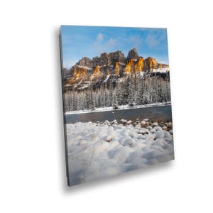 May include: A snowy landscape with a mountain range in the background. The mountains are a golden colour and the snow is white. There is a river in the foreground and some trees in the middle ground.