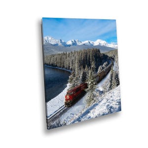 May include: A red train engine pulls a line of boxcars along a snowy track through a wintery landscape. The train is traveling through a forest of snow-covered trees and past a river. The mountains in the background are also covered in snow.