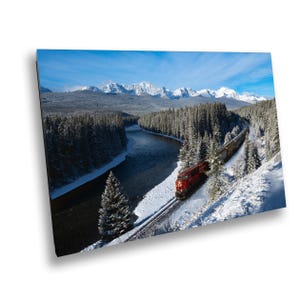 May include: A red train travels through a snowy mountain landscape. The train is moving along a track that winds through a forest and alongside a river. The mountains in the background are covered in snow and the sky is blue.