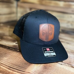 May include: Black trucker hat with a brown leather patch featuring an American flag and a skull design. The patch is stitched onto the front of the hat. The hat is adjustable and has a mesh back.