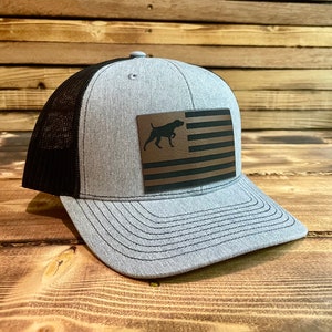 May include: A gray and black trucker hat with a brown leather patch featuring an American flag with a silhouette of a dog.
