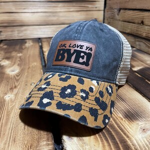 May include: A charcoal grey trucker hat featuring a tan leopard print brim and a rectangular brown leather patch on the front. The patch is embossed with the black text "OK, LOVE YA BYE!" and the hat includes a beige mesh back panel.