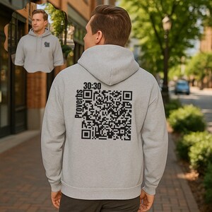 Puede incluir: Una sudadera con capucha gris claro con mangas largas. La parte trasera de la sudadera presenta un gran código QR negro con el texto "30:30" encima y la palabra "Proverbios" a la izquierda. Una imagen más pequeña de la sudadera está en la esquina superior izquierda.