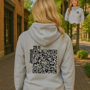 May include: A pale grey hoodie featuring a black QR code and the text "29:11 Jeremiah" on the back. The hoodie has a hood and long sleeves. The image shows a person wearing the hoodie.