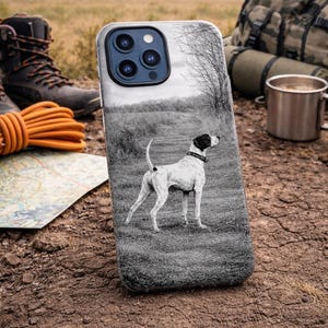 May include: A black and white phone case featuring a dog standing in a field. The case has a black border and a picture of a dog with a black collar. The background includes a map, boots, and camping gear.