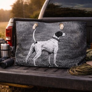 May include: A large tote bag featuring a black and white illustration of a hunting dog standing in a field. The bag has tan leather handles and is resting in the back of a truck. Other items include binoculars, a thermos, and a hat.