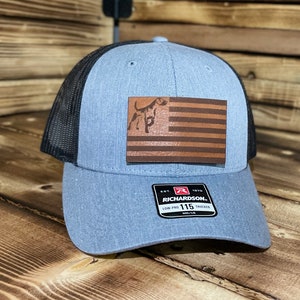 Puede incluir: Gorra de camionero azul y negra con un parche de madera de la bandera americana con la silueta de un perro. La gorra tiene la parte trasera de malla y una visera curva. El parche es marrón.