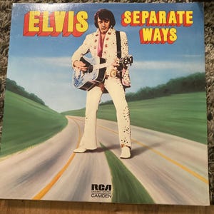 May include: Album cover for "Elvis Separate Ways" featuring Elvis Presley in a white jumpsuit with floral accents, playing a guitar. The album title is in bold yellow and red letters against a blue sky and road background. The RCA Camden logo is visible.